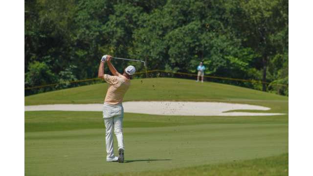 A man in a tan shirt, white pants, and a white hat swings a golf club overhead, sending the ball over a sand trap toward the hole.