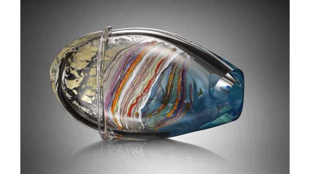 A sculpture that looks like an oval vase turned on its side. It looks like multiple layers of glass, with the innermost layer formed of colorful bands (pink, lavender, red, orange, green, brown, white, blue, and more). The next layer looks clear or blue. The outermost layer appears clear with silver stripes and a golden leopard-like pattern.