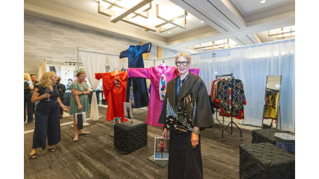 An art booth at the Texas Fine Craft Show features a prominent display of three East Asian-inspired clothing designs, one bright red, one blue, one hot pink. SuzKaz, a woman with short-cropped blonde hair, thick-rimmed glasses, and a thick animal-skin shawl, stands with her work as guests admire her work.