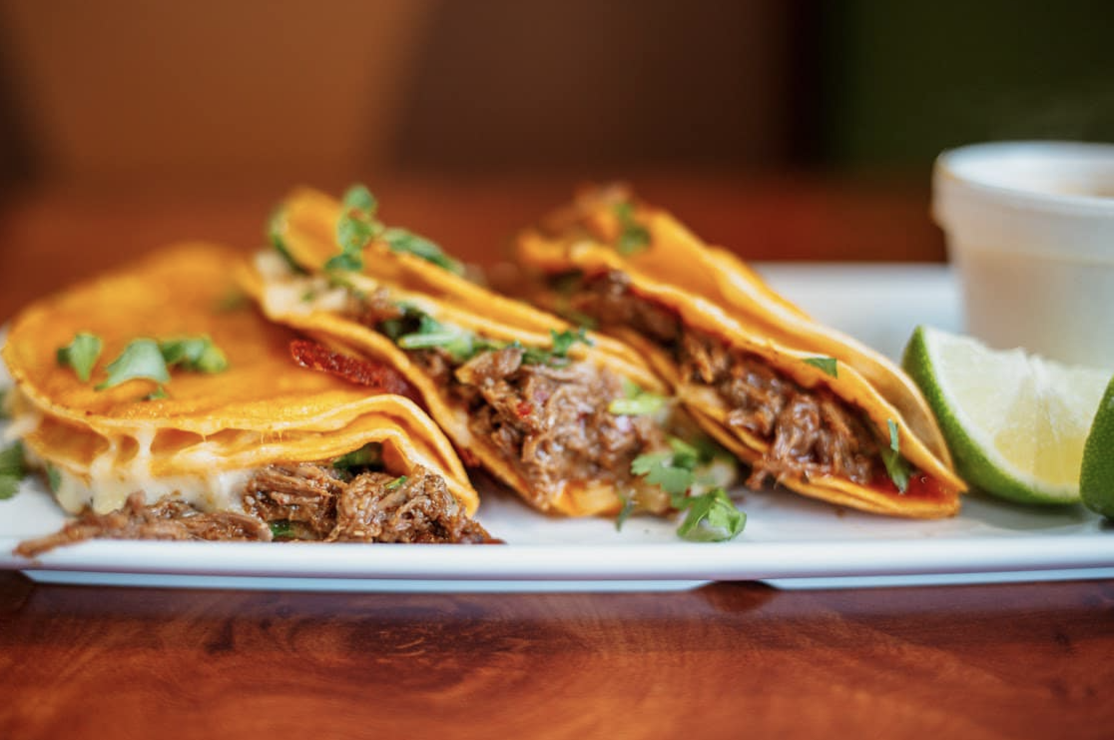 A plate of tacos with meat, cheese, and cilantro, accompanied by a lime wedge and a small cup of sauce.