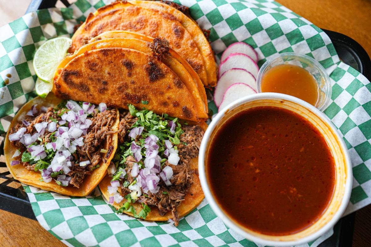 Crispy birria tacos topped with onion and cilantro next to a cup of consomme at Red Tacos