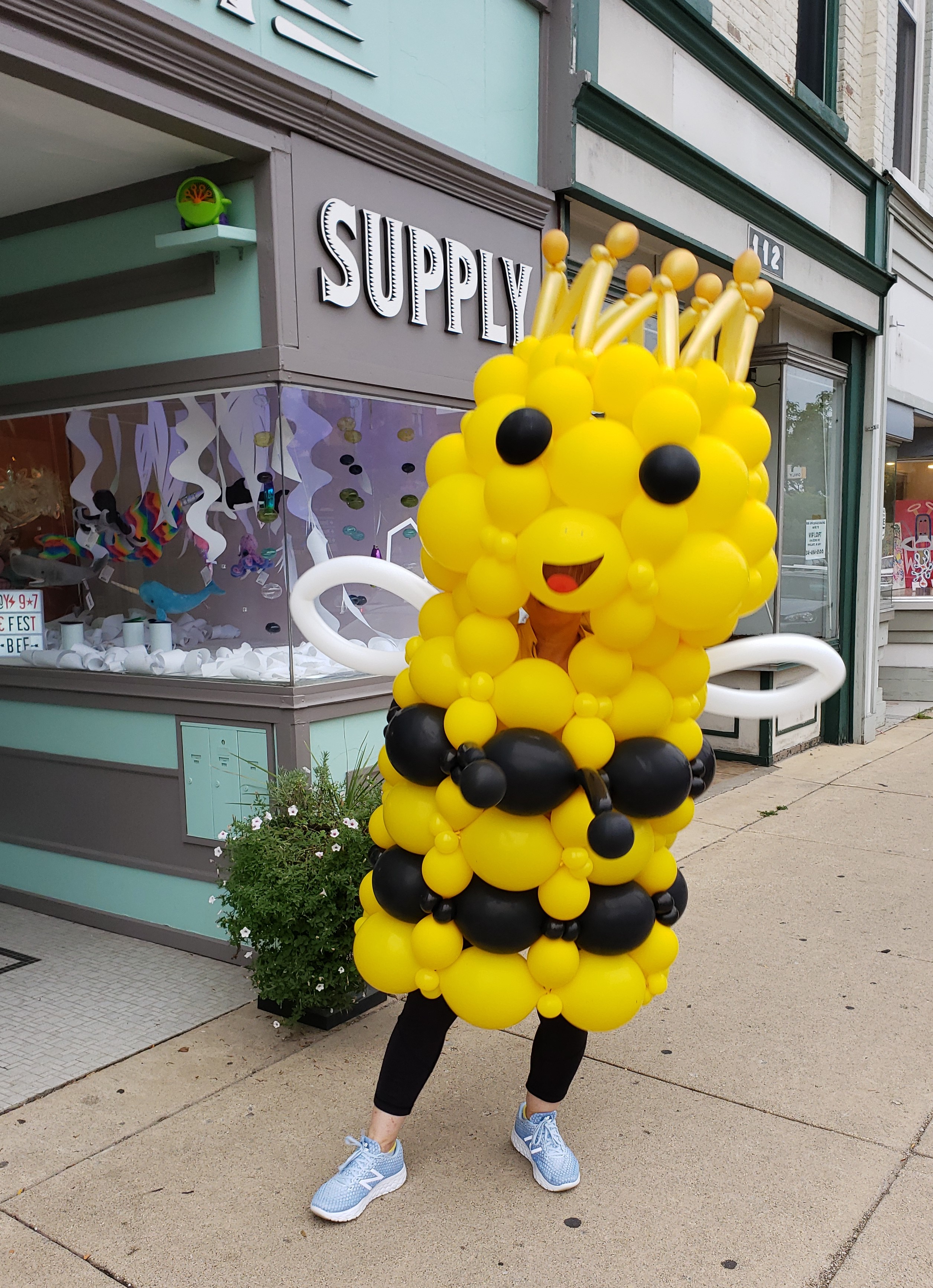 A person wears the Queen Bizzy Bee costume, a yellow and black balloon sculpture of a smiling queen honeybee.