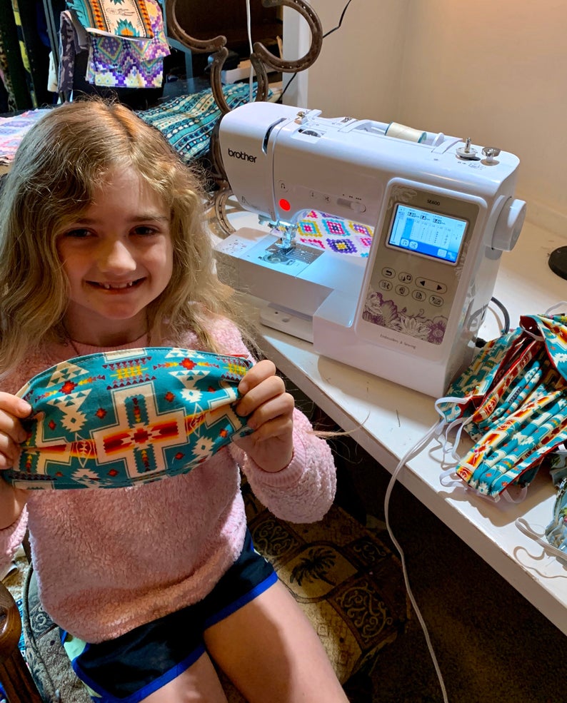 Young girl sits next to a sewing machine and displays a mask she made from Native American themed fabric.
