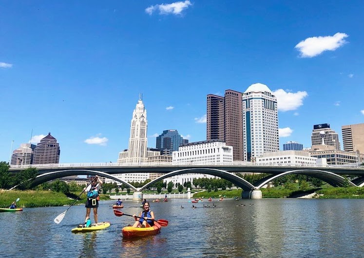 Kayakers in Scioto River with Scioto Mile and city skyline in background under blue sky