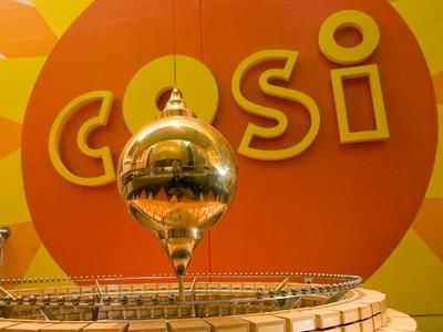 A Foucalt Pendulum swinging in front of a COSI sign