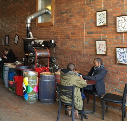 Two people enjoying coffee next to giant coffeemakin apparatus, brick walls lined with artwork inside Upper Cup Coffee