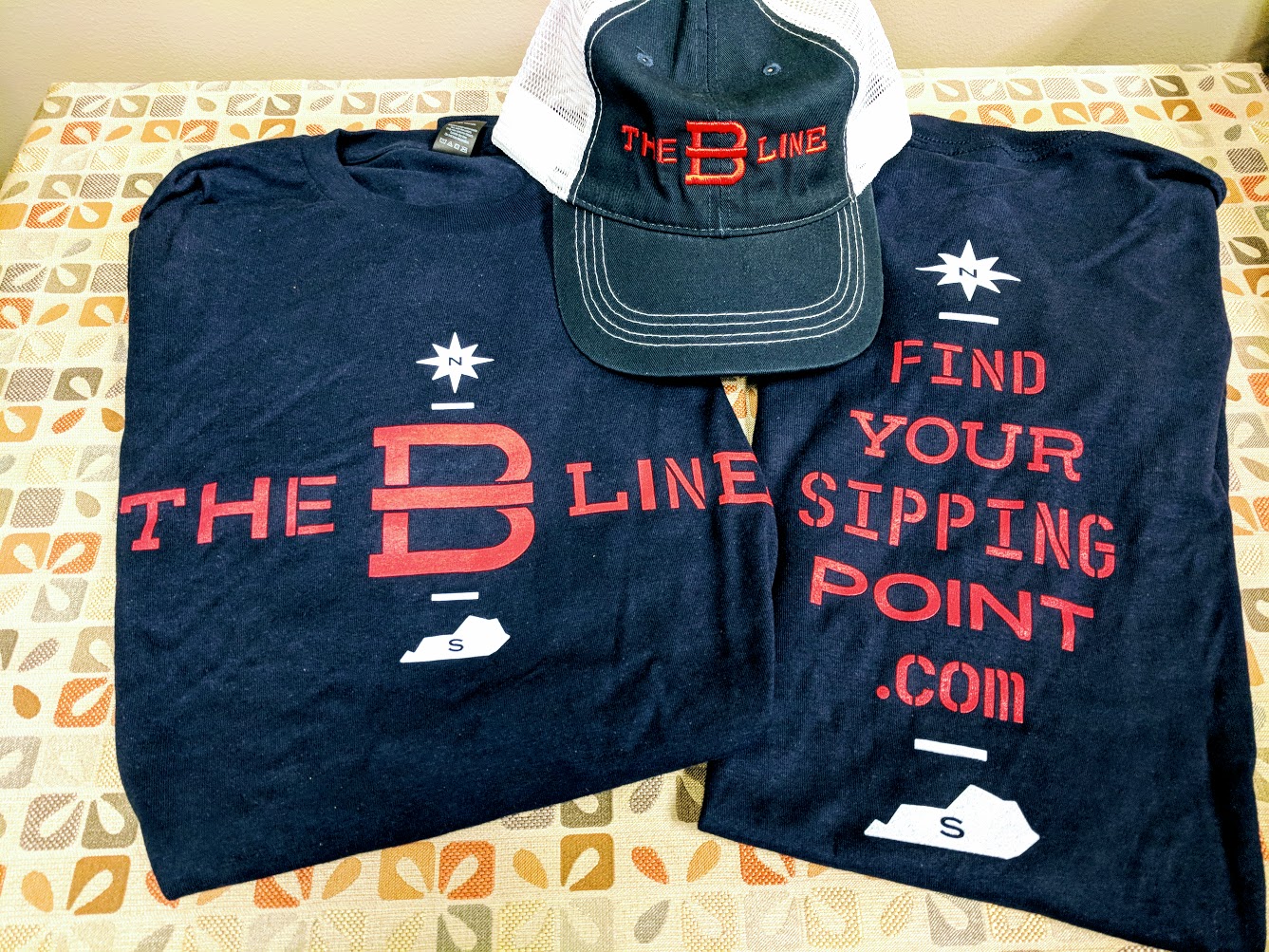 Some of the free bourbon swag people get for completing the B-Line Bourbon trail in NKY. Blue, white and red t-shirts and a hat that have the words The B-Line, Find Your Sipping Point