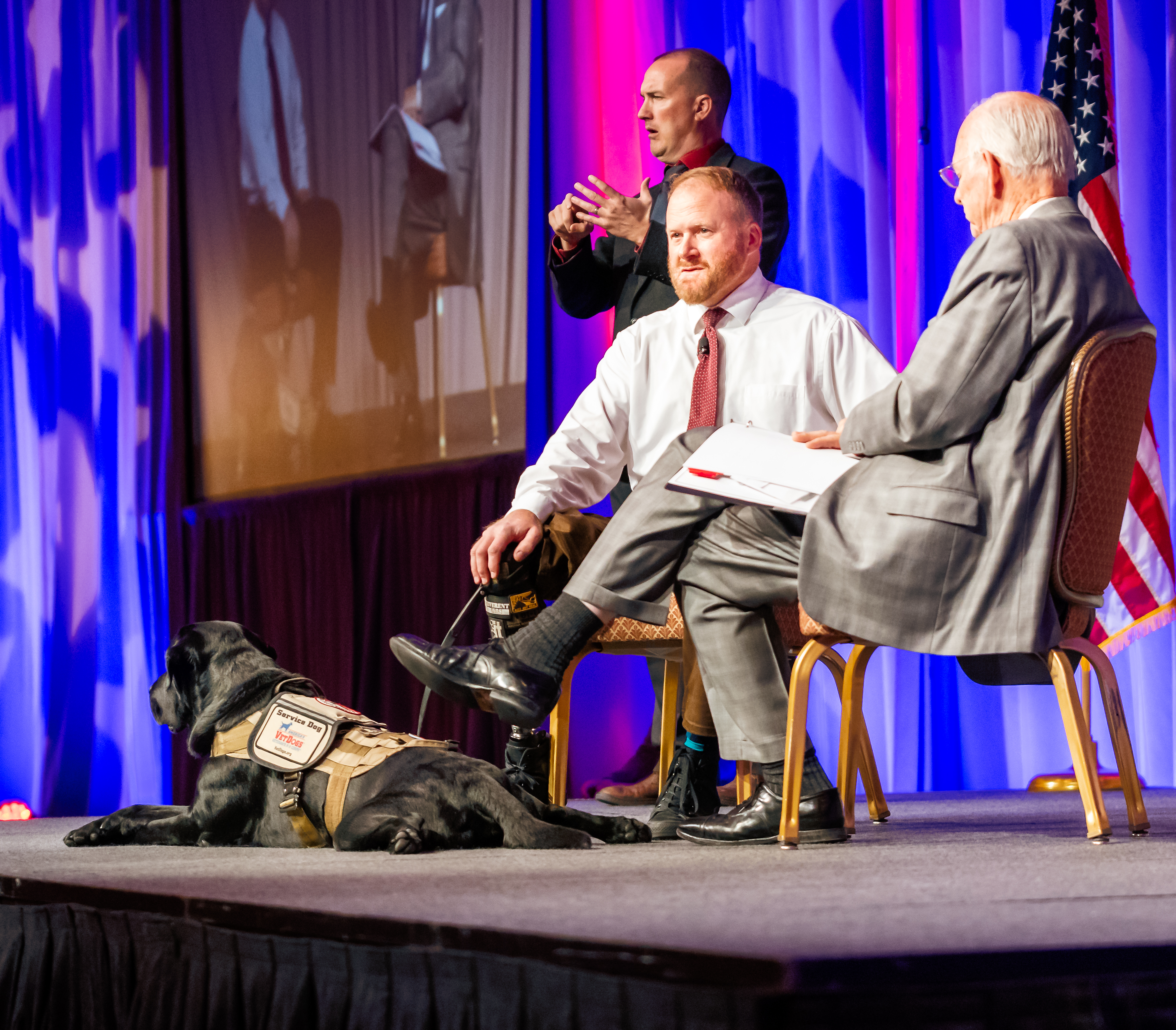 Two People speaking on a stage with a service dog laying down next to them