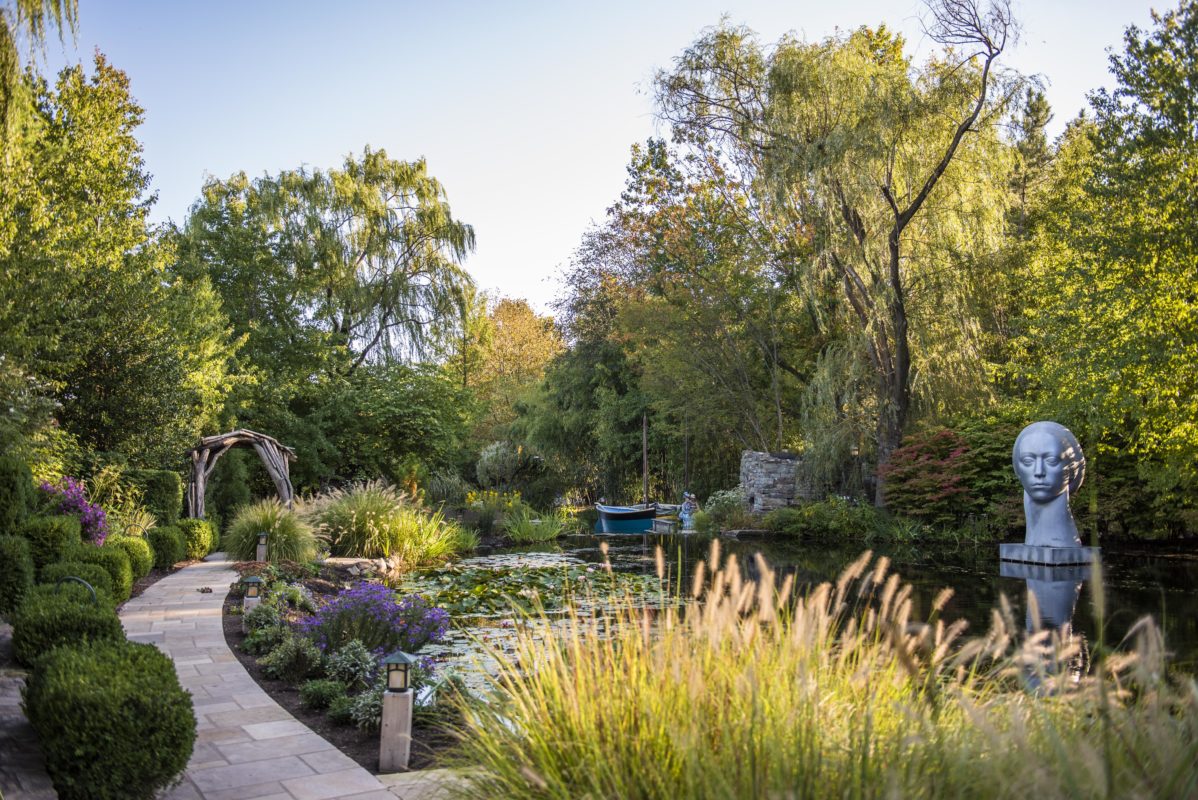 This is a photo of a pond, surrounded by trees with a walkway to the left of he pond. Surrounding a pond is gorgeous landscaping such as bushes and plants.