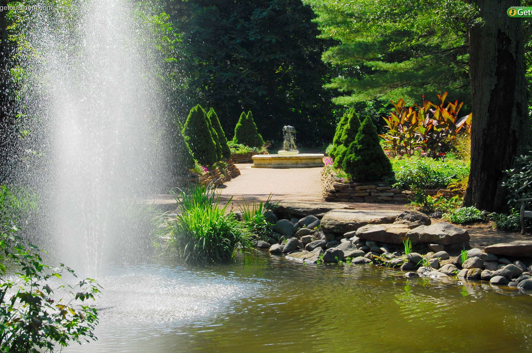 A large fountain sprays water over a flower bed. A path behind the flowers leads up to a sculpture.