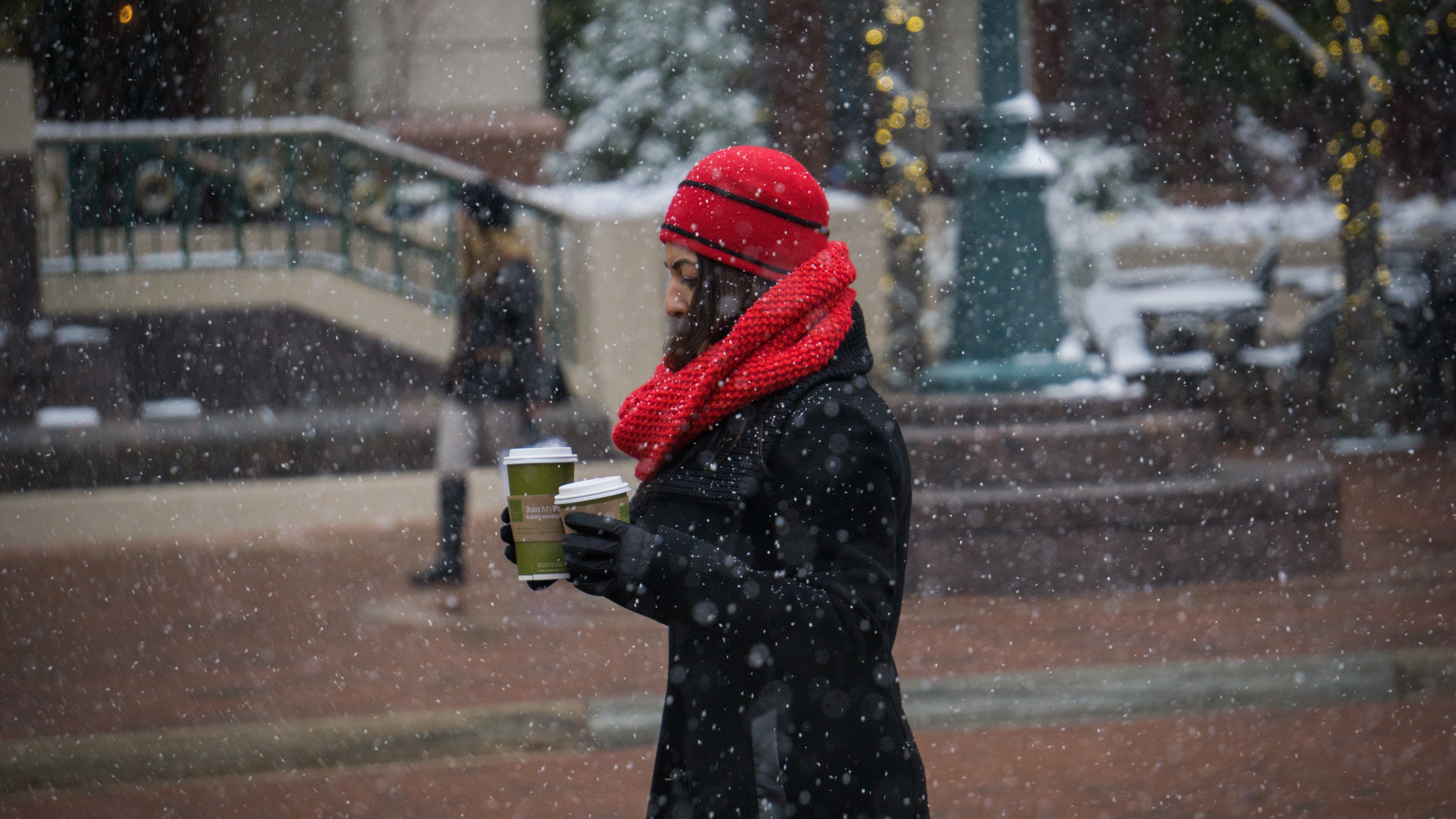 Woman in a red winter hat and scarf walking in the snow holding two cups of coffee.