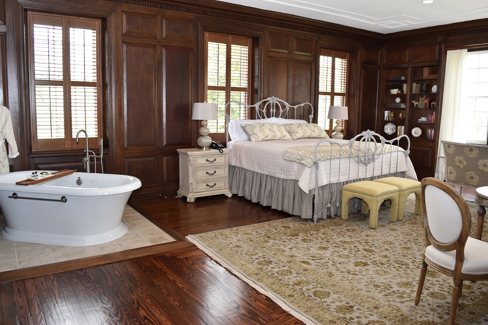 a room at an inn, the room has wood paneling, a bathtub, a side-table, a bed with a white comforter, foot stools and a chair with hardwood floors and a white and gold floral rug.