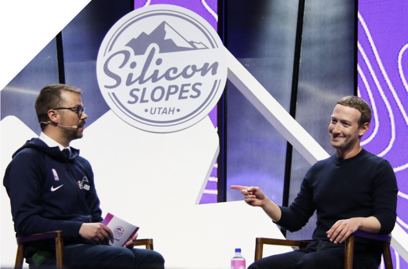 Two seated speakers on stage with teal blue curtains behind them and the projected image of the Silicon Slopes logo on the curtains