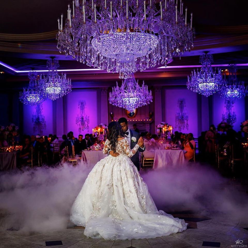 Bride and groom dancing under chandeliers in dark room lit in purple lights with mist on the ground