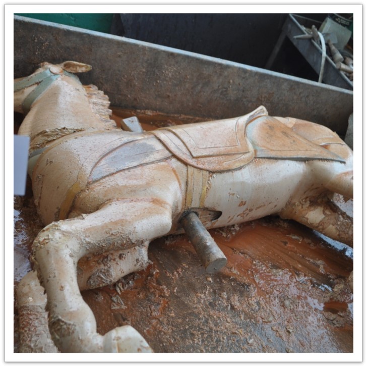 Carousel horse laying on its side on a workshop floor, in the early stages of having old finish stripped.