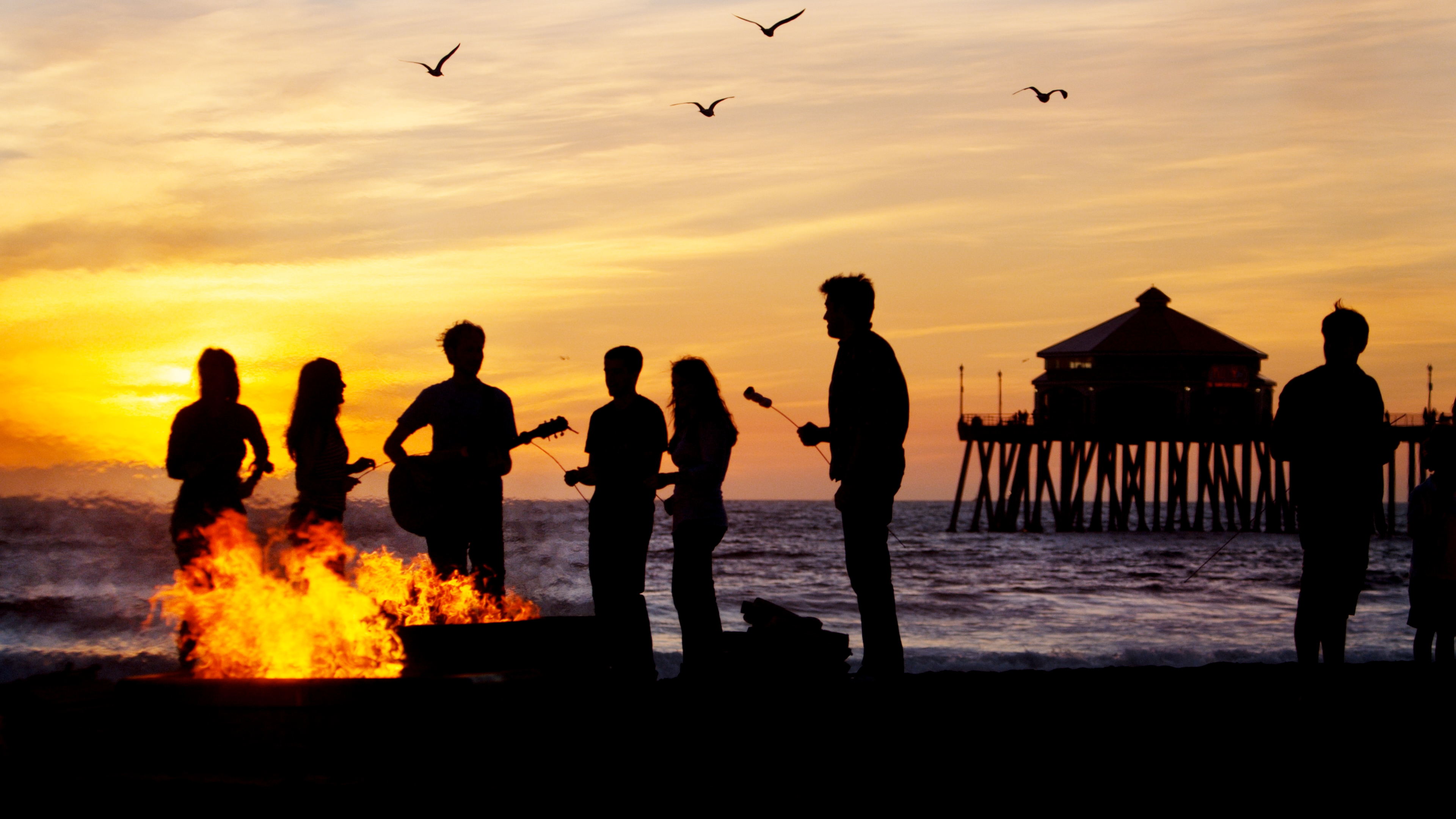 Friends on the beach at sunset playing music and roasting marshmallows by a bonfire in Huntington Beach 
