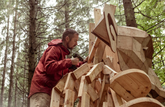 Thomas Dambo works on a large wooden troll sculpture among trees.
