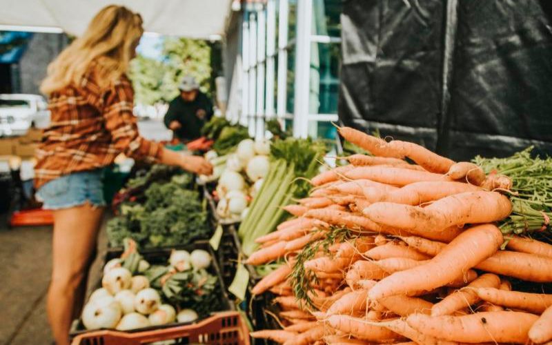 A woman browsing fresh vegetables at an outdoor vendor stall at the Vancouver Farmers Market, with carrots in the foreground