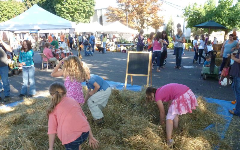 Children playing in a hay bale area at the Camas Farmers Market with vendor tents and families in the background