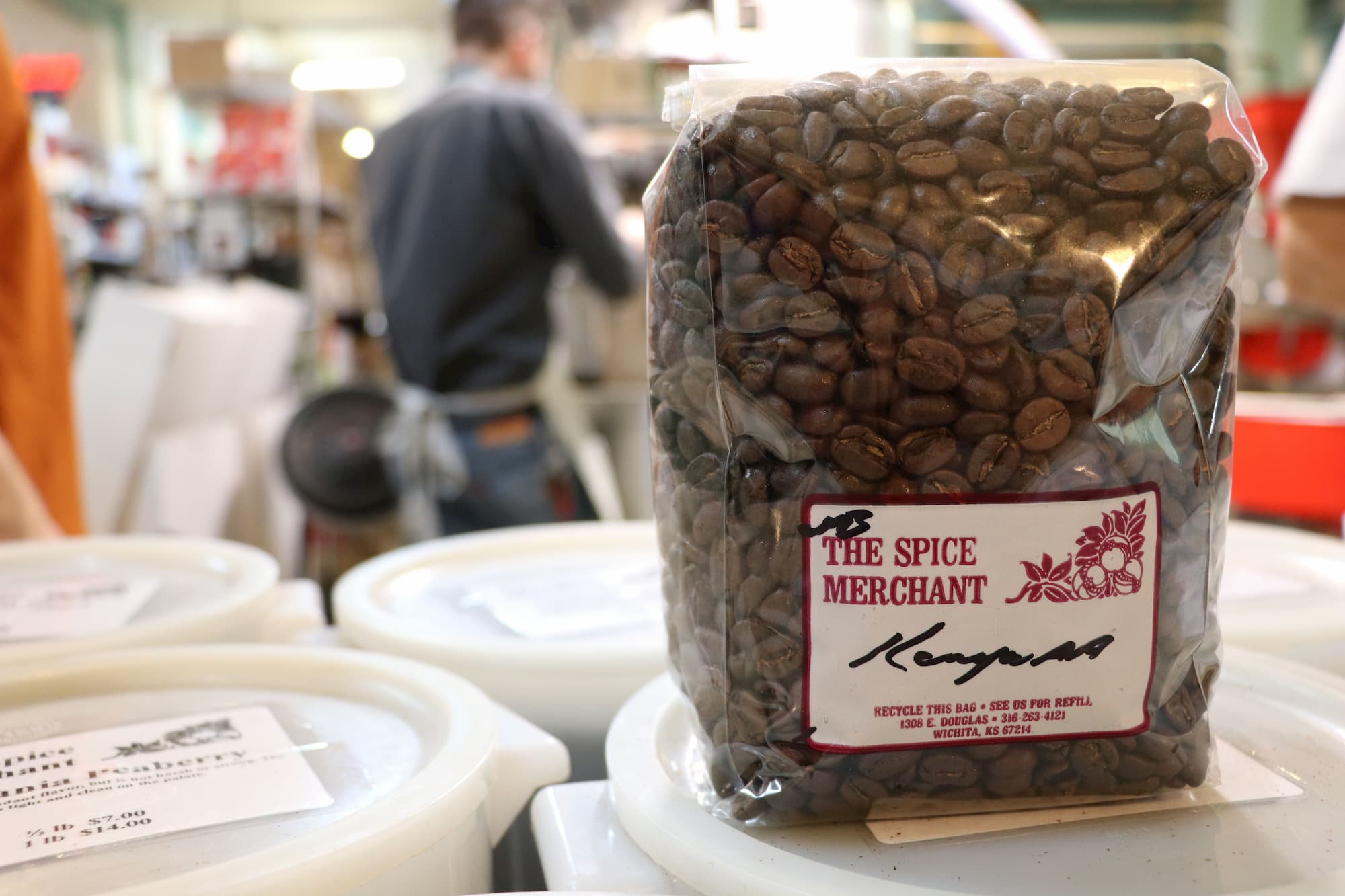 A bag of Spice Merchant coffee beans is in the foreground, while a worker rings someone up in the background