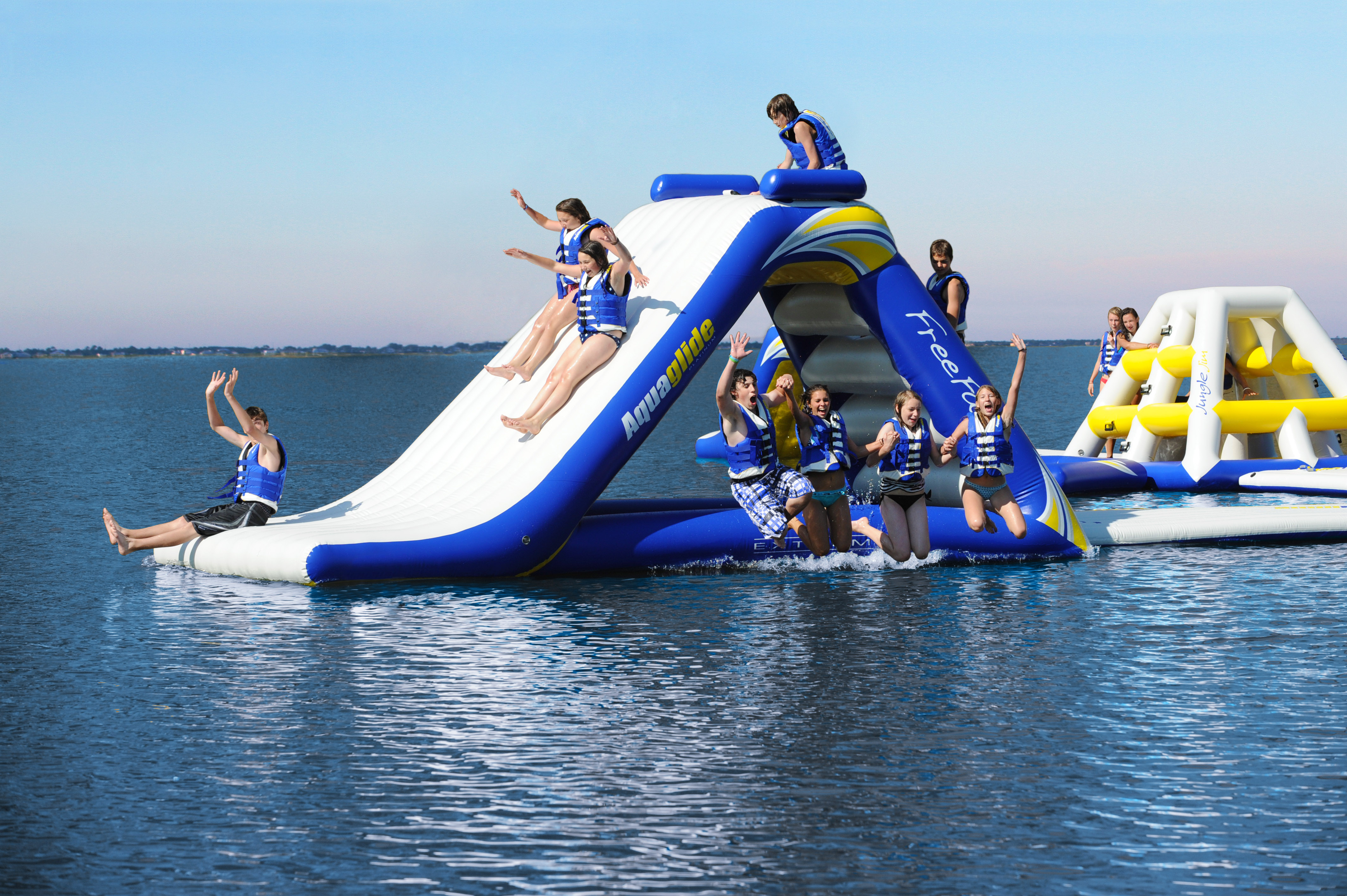 Kids slide down an inflatable slide in a lake while other kids jump into the lake off the side