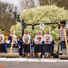 A group of young women in traditional Dutch costumes with white bonnets, floral aprons, and wooden clogs hold bouquets of pink, red, and yellow tulips during the ceremonial street scrubbing to kick off the Albany Tulip Festival, with brooms and buckets visible in the foreground.