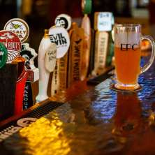 A row of colorful craft beer taps lines a polished bar, with a glass of golden beer labeled “EPC” resting on a coaster in the foreground under warm bar lighting at the Excelsior Pub in Albany, New York.