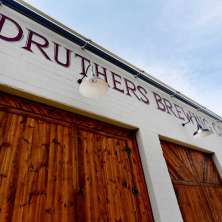 Exterior view of Druthers Brewing Company in Albany with a white brick facade, large wooden barn-style doors, and the brewery’s name painted across the building under mounted exterior lights.
