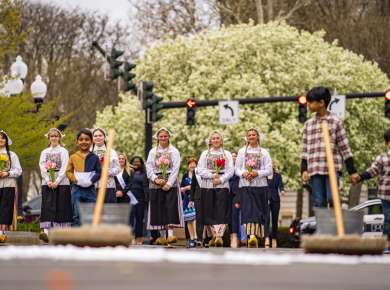 A group of young women in traditional Dutch costumes with white bonnets, floral aprons, and wooden clogs hold bouquets of pink, red, and yellow tulips during the ceremonial street scrubbing to kick off the Albany Tulip Festival, with brooms and buckets visible in the foreground.