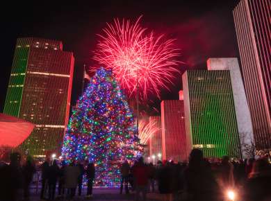 A large Christmas tree covered in colorful lights glows in the center of Empire State Plaza at night as a crowd looks on, with red and green–lit office towers and bright red fireworks bursting in the sky overhead.
