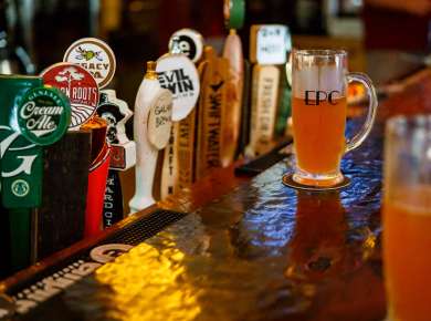 A row of colorful craft beer taps lines a polished bar, with a glass of golden beer labeled “EPC” resting on a coaster in the foreground under warm bar lighting at the Excelsior Pub in Albany, New York.