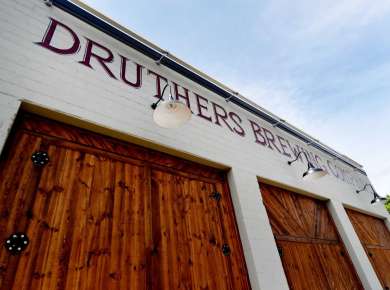 Exterior view of Druthers Brewing Company in Albany with a white brick facade, large wooden barn-style doors, and the brewery’s name painted across the building under mounted exterior lights.