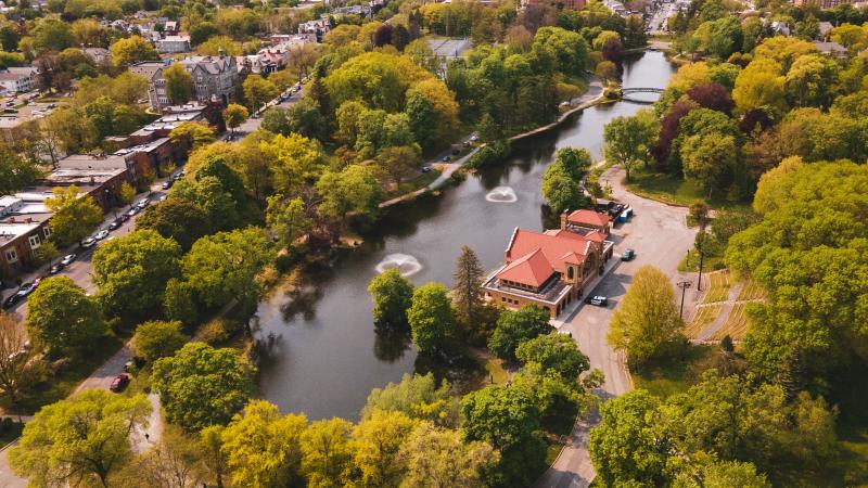 Aerial view of Washington Park’s lake lined with lush spring trees, with two small fountain sprays in the water, a red-roof lakehouse/boathouse on the shoreline, and a footbridge in the distance near surrounding neighborhoods.