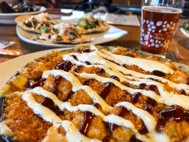 Close-up of a baked mac and cheese topped with crispy breadcrumbs, drizzled with barbecue sauce and a creamy white sauce, served on a plate at a wooden table with flatbread appetizers and a pint of beer blurred in the background