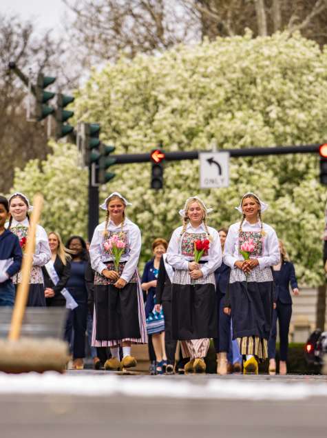 A group of young women in traditional Dutch costumes with white bonnets, floral aprons, and wooden clogs hold bouquets of pink, red, and yellow tulips during the ceremonial street scrubbing to kick off the Albany Tulip Festival, with brooms and buckets visible in the foreground.