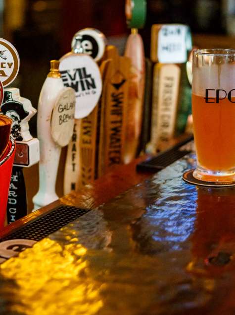 A row of colorful craft beer taps lines a polished bar, with a glass of golden beer labeled “EPC” resting on a coaster in the foreground under warm bar lighting at the Excelsior Pub in Albany, New York.