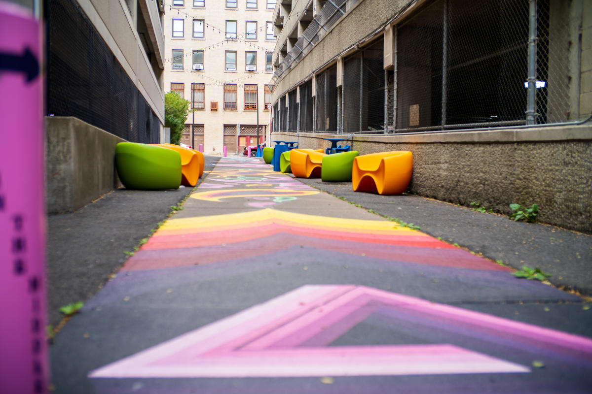 Colorful street art covers the pavement of a narrow urban alleyway in Albany, NY, with vibrant chevron and geometric patterns leading toward a building at the end. Pairs of green and orange molded chairs line both sides of the alley, with string lights overhead.