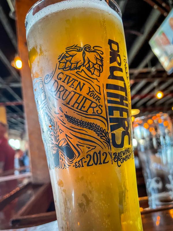 Close-up of a tall pint glass filled with golden beer and a foamy head, featuring the Druthers Brewing Company logo and “Given Your Druthers, Est. 2012,” with warm bar lighting and taps blurred in the background.