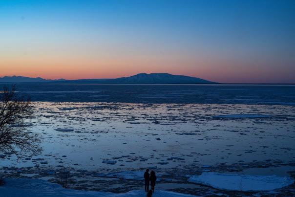 People stand at the edge of Cook Inlet with Mount Susitna across the water at dusk.
