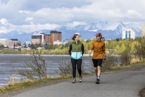 Two people walking on the Tony Knowles Coastal Trail with downtown Anchorage in the background.