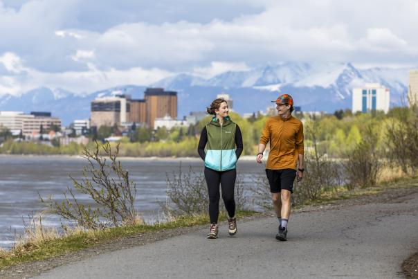 Two people walking on the Tony Knowles Coastal Trail with downtown Anchorage in the background.