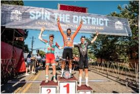 Winners of a men's cycling event stand with raised arms on a podium in front of an overhanging banner that reads "Spin the District"