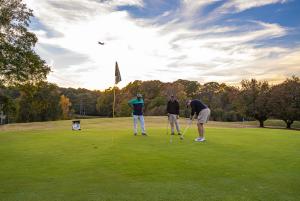 Three men play on a golf course with an airplane flying through the cloud marbled sky in the background.