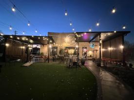 Green outdoor space in front of Paper Plane restaurant at dusk with string lights illuminated.