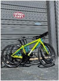 Profile shot of three brightly colored hybrid bikes in a bike rack in front of a gray garage door with the Outback Bike's red and white oval logo.