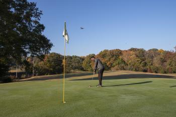 Man putts on golf course as plane flies overhead.