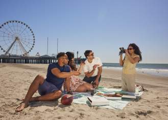 Young couples sit and enjoy pizza on the Atlantic City beach with Steel Pier in background. Woman kneeling holds a camera to snap a picture.