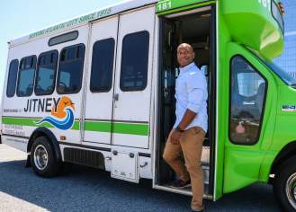 A person stands next to a green and white jitney bus, showcasing its service for Atlantic City, with a clear sky in the background.