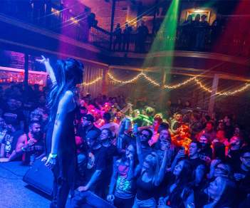 A performer on stage with arms raised as crowds watch bathed in rainbow colored lighting at Anchor Rock Club in Atlantic City, NJ.