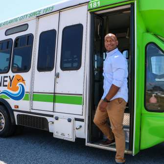 A person stands next to a green and white jitney bus, showcasing its service for Atlantic City, with a clear sky in the background.